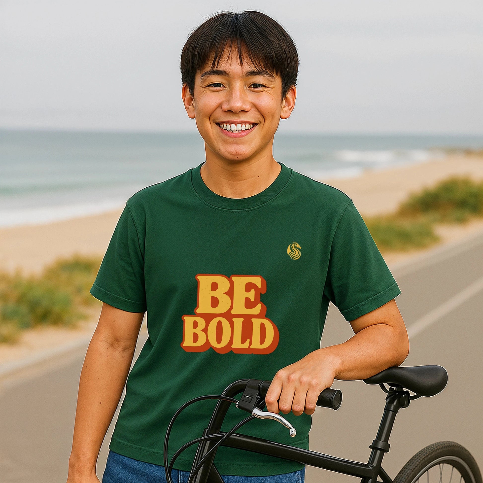 Be Bold Youth 100% Organic Cotton T-Shirt green “BE BOLD” graphic worn by teen outdoors by beach holding bicycle