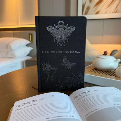 Diario de Gratitud Negro con Silver Lining – 120 páginas de I am Thankful For sobre una mesa, tapa negra con diseño plateado, páginas abiertas