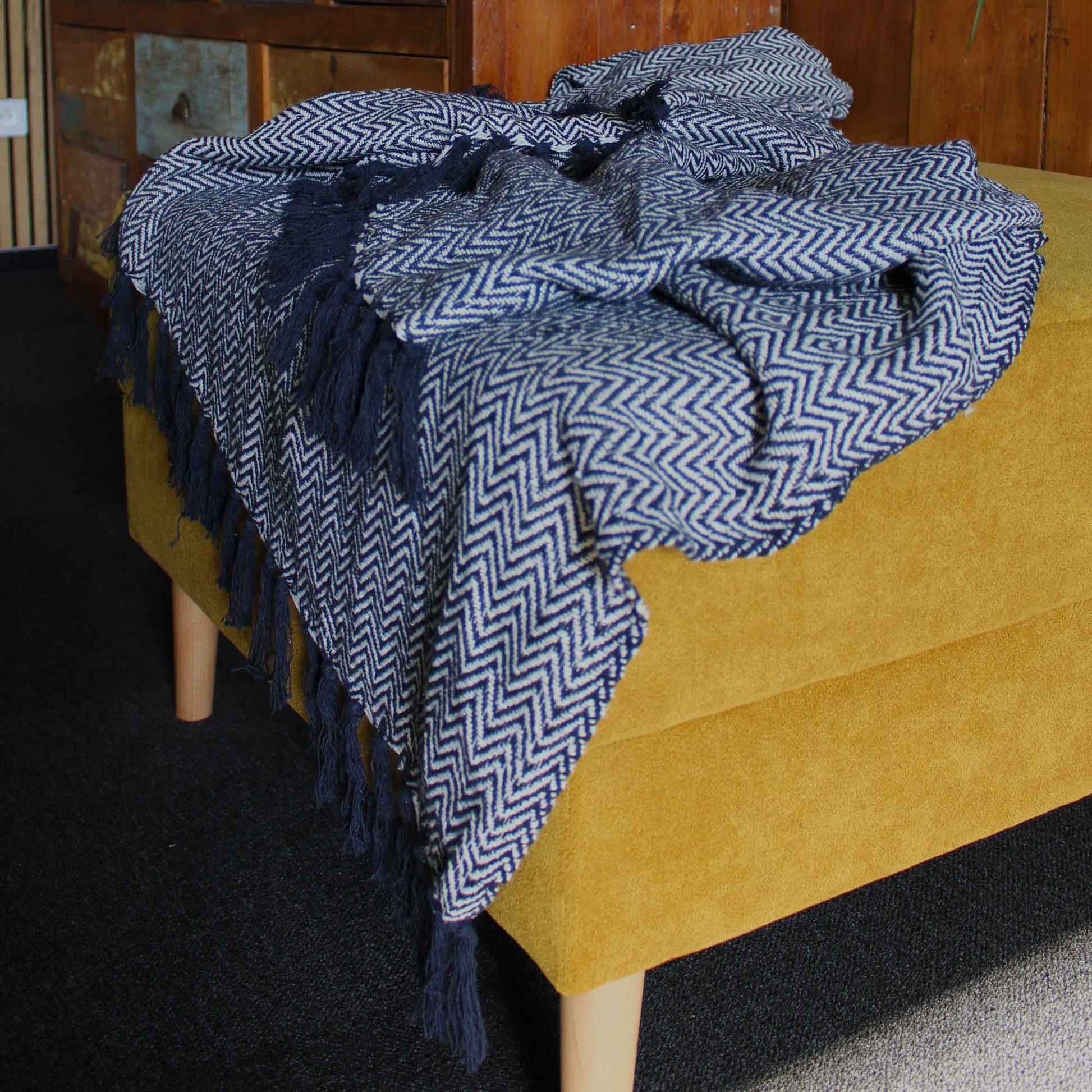 Boho Comfort-pläd – Marinblå handvävd pläd i bomull med marinblått sicksackmönster och fransar, draperad över en senapsgul ottoman