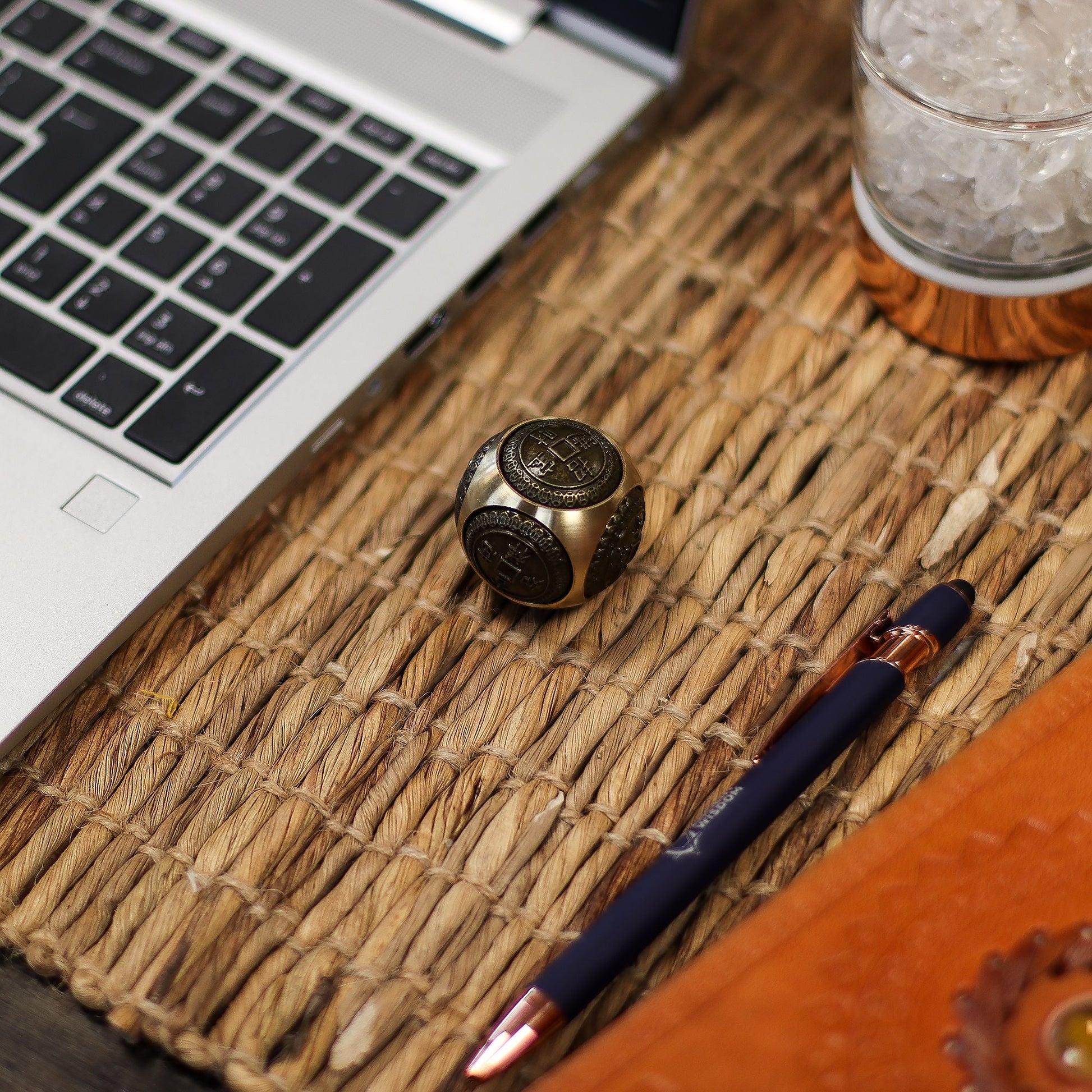 Alt Tag: Brass Ancient Fortune Spinner – Rounded 3.5 CM de latón con motivos de moneda china sobre escritorio junto a laptop y bolígrafo