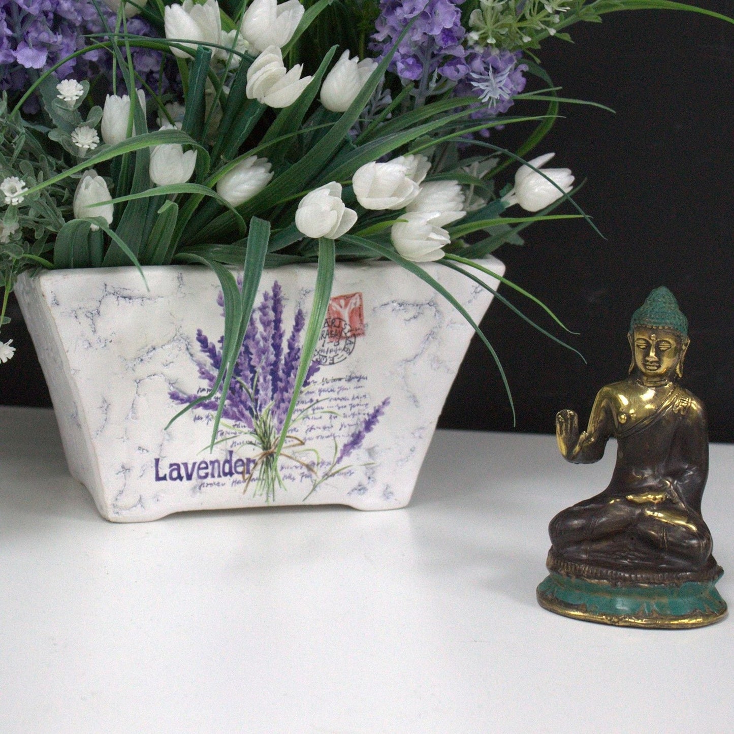 Alt Tag: Brass Buddha Feng Shui Statue – 6x4 CM de latón en postura de meditación junto a maceta decorativa con flores blancas y lavanda