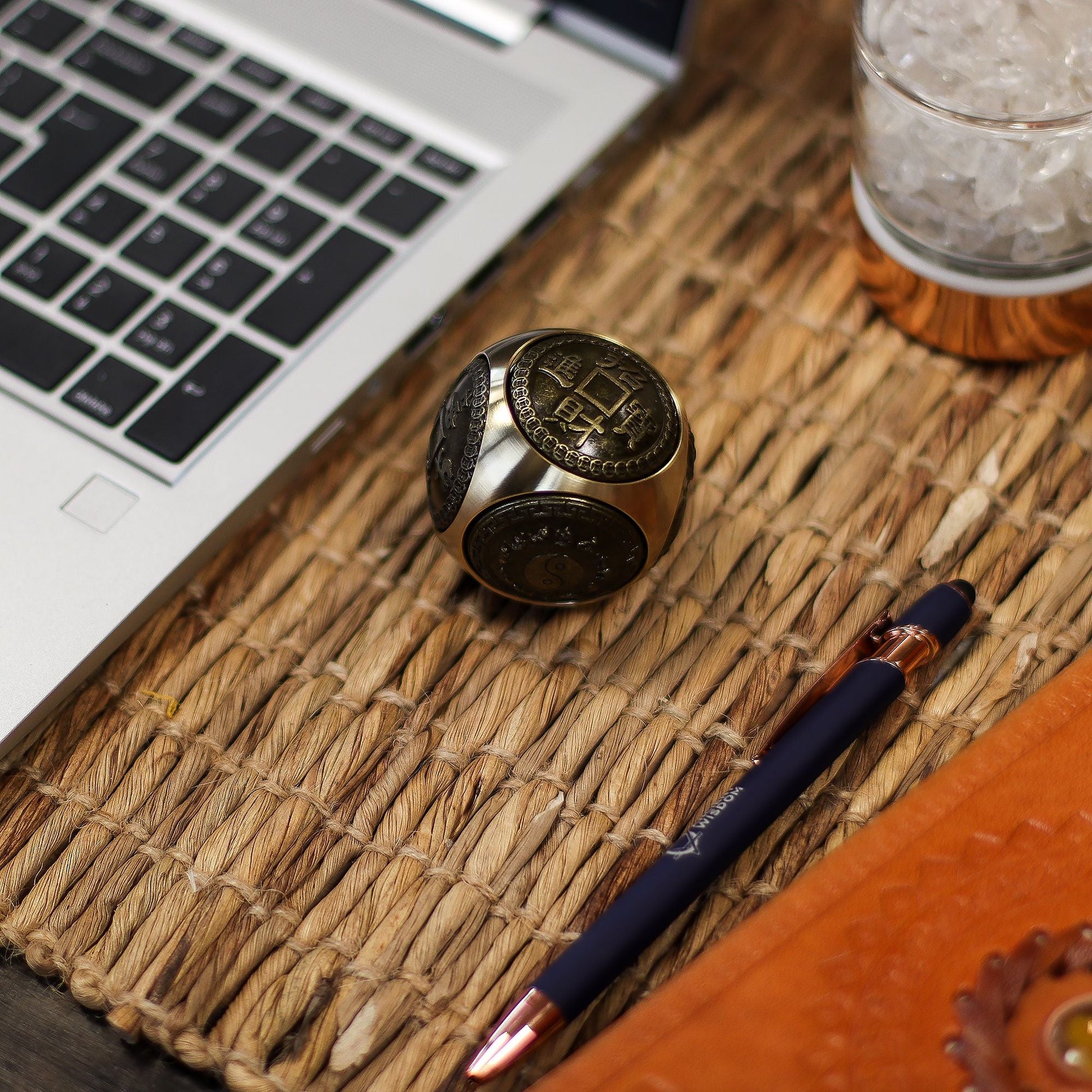 Brass Feng Shui Wealth Spinner – 5.5 CM on woven desk mat beside laptop, pen and glass, showing engraved Chinese coin design