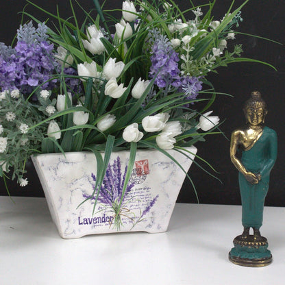 Stehende Buddha-Statue aus Messing – 17x4 CM neben einem weißen und lavendelfarbenen Blumenarrangement in einem Keramiktopf, Messingfigur mit grünem Gewand
