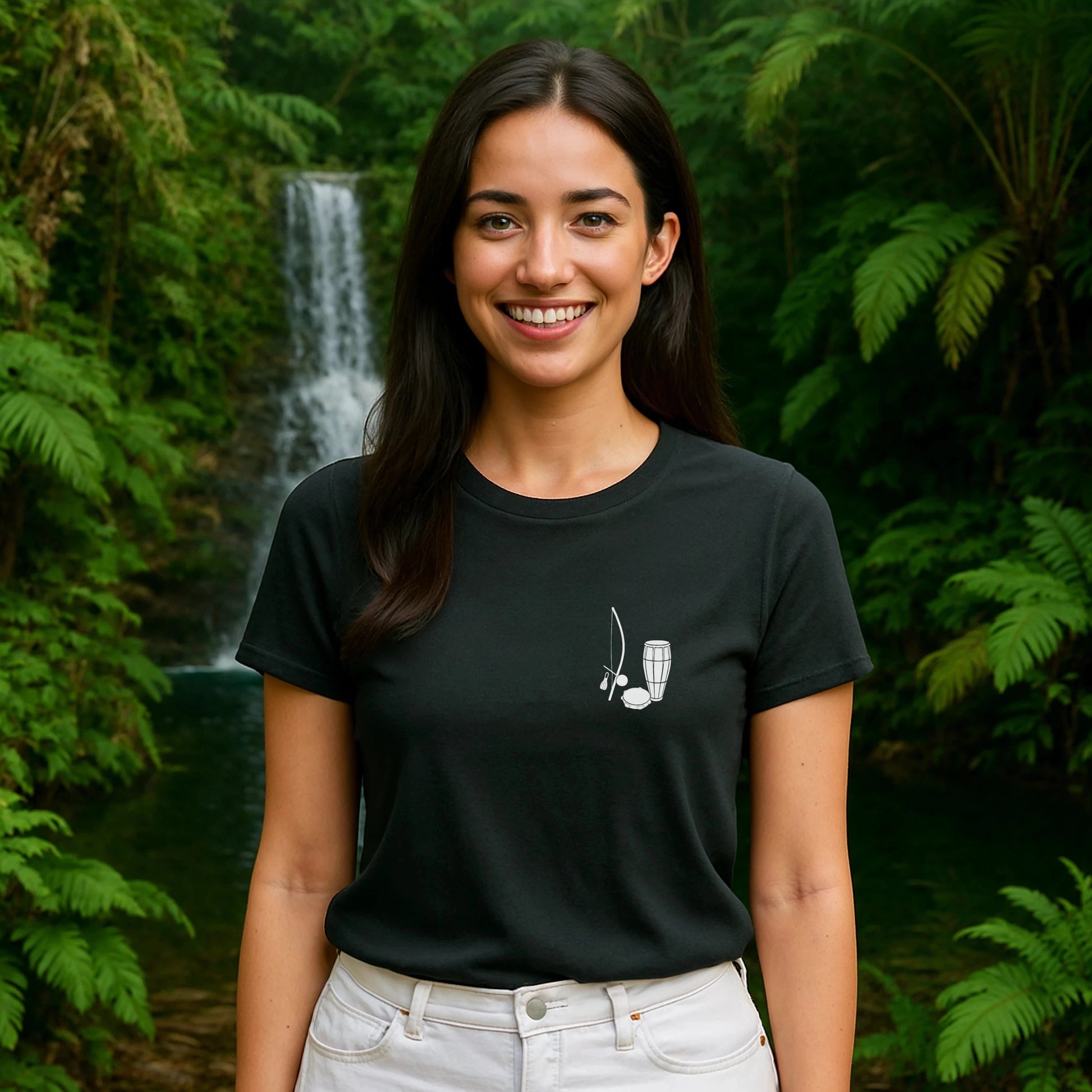 Capoeira Music Sustainable Unisex Tee on model, black t-shirt with berimbau graphic, standing outdoors by lush waterfall