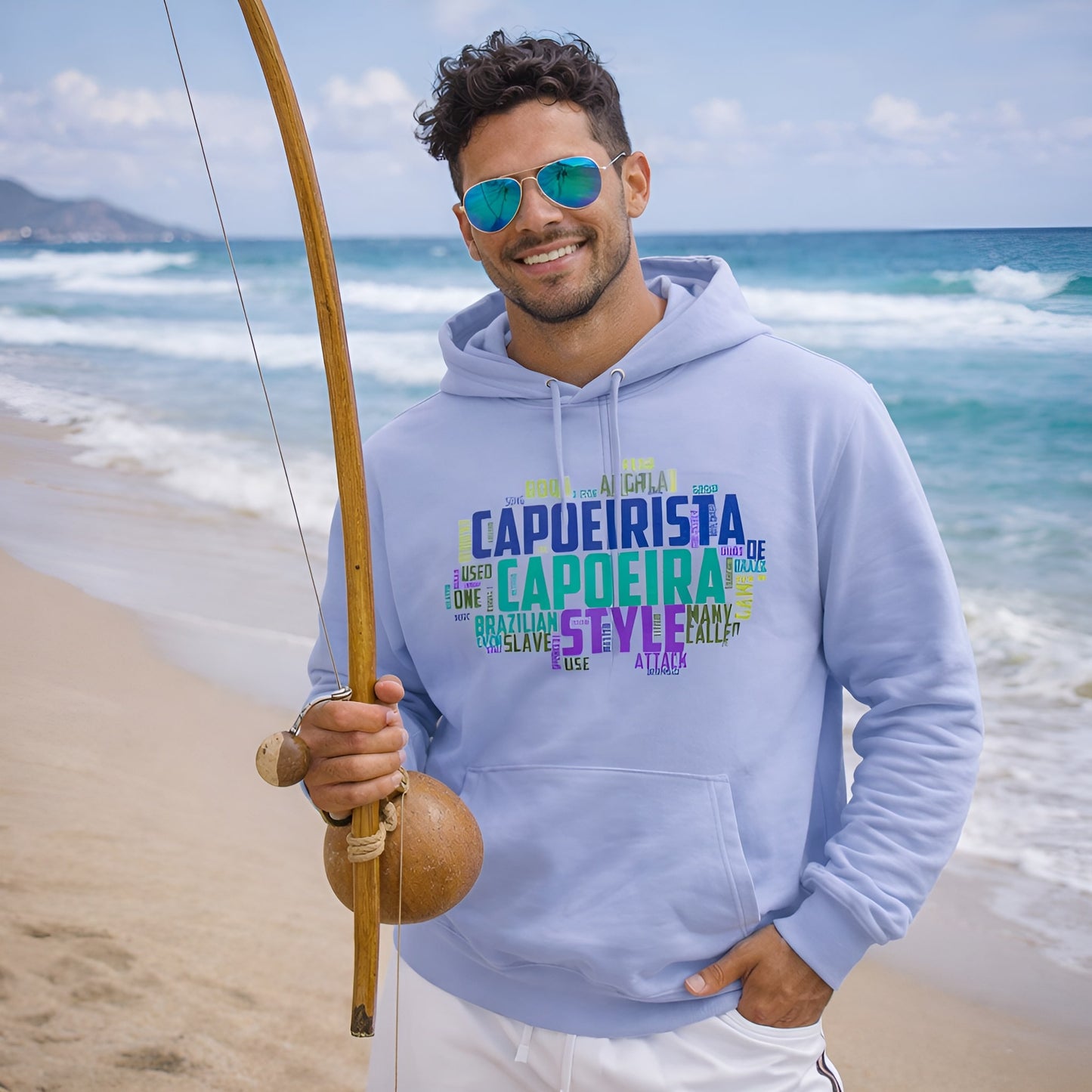 Capoeira Sustainable Unisex Hoodie in light blue organic cotton with capoeira word art print, worn on beach with berimbau