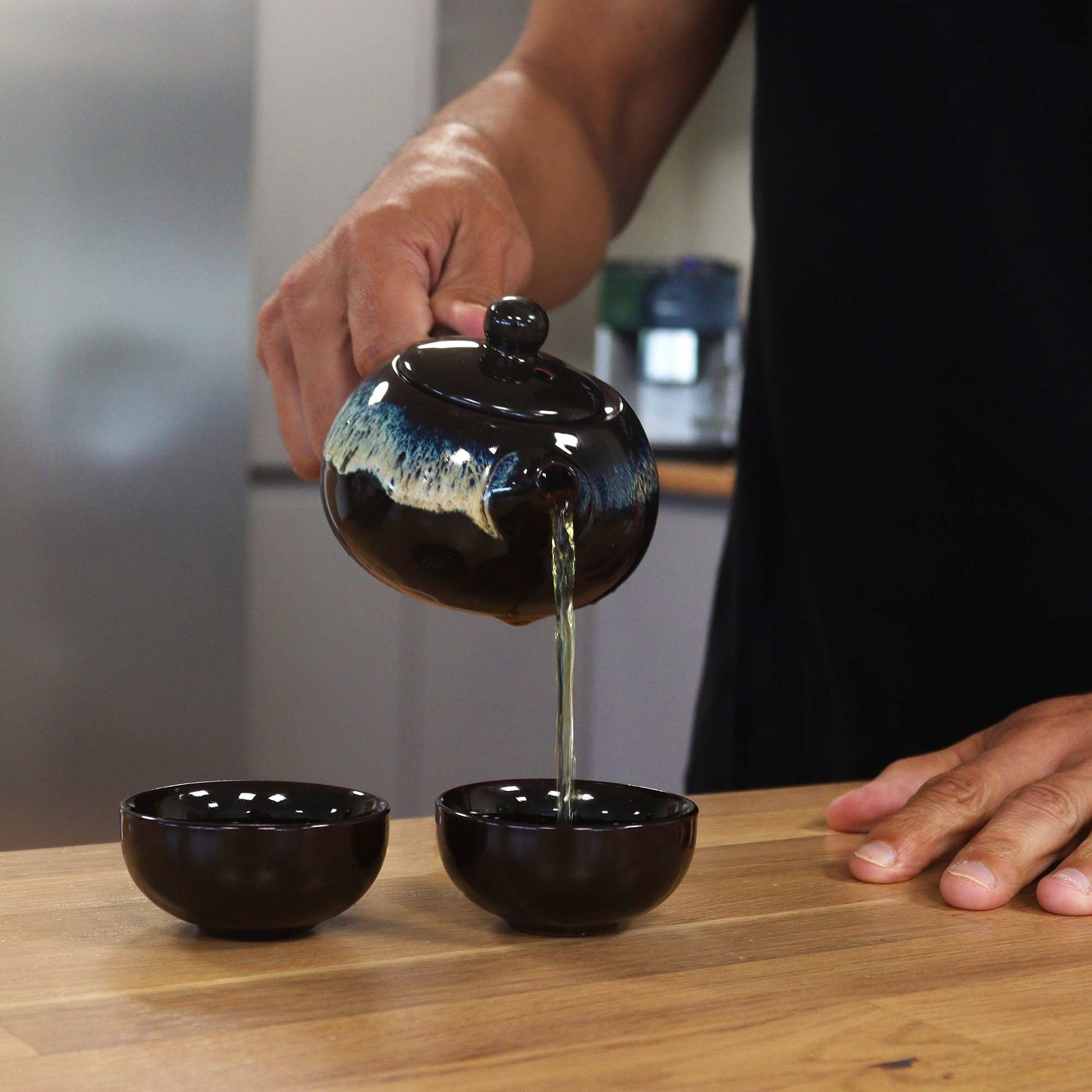 Ceramic Herbal Teapot Set – 19x12.5x9 CM black glazed ceramic teapot pouring tea into small cups on wooden table