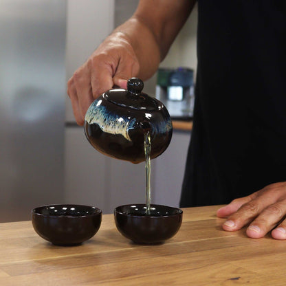 Ceramic Herbal Teapot Set – 19x12.5x9 CM black glazed ceramic teapot pouring tea into small cups on wooden table