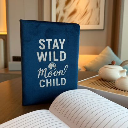 Cuaderno de bienestar de terciopelo azul oscuro – 80 páginas con tapa de terciopelo con la frase “Stay Wild Moon Child”, colocado junto a páginas rayadas abiertas
