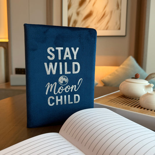 Dark Blue Velvet Wellness Notebook – 80 Pages with velvet cover reading “Stay Wild Moon Child”, standing beside open lined pages