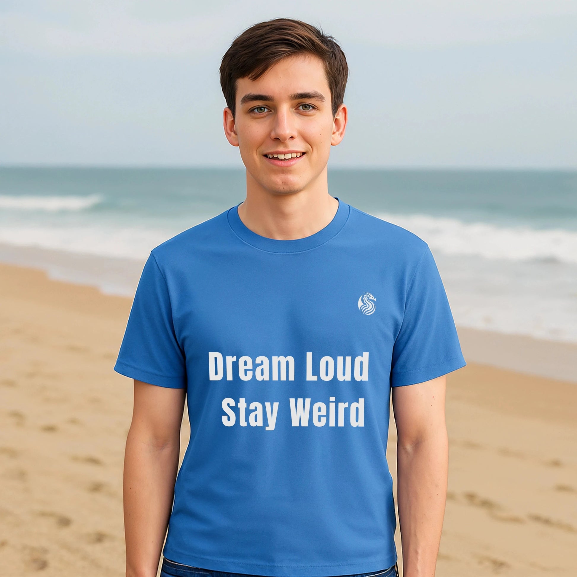 Dream Loud. Stay Weird % Drix Sustainable Unisex Tee camiseta azul con texto “Dream Loud Stay Weird” en modelo frente al mar