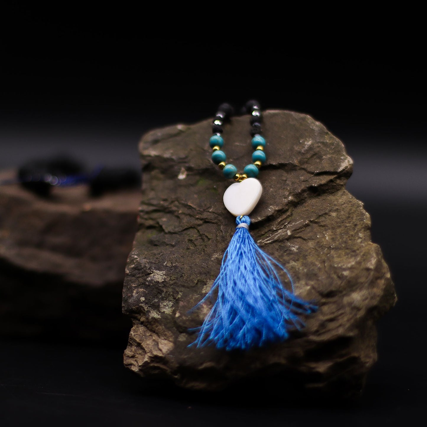 Essence of the Volcano Necklace – Calming Influence with lava stone beads, turquoise accents, white heart pendant and blue tassel on rock