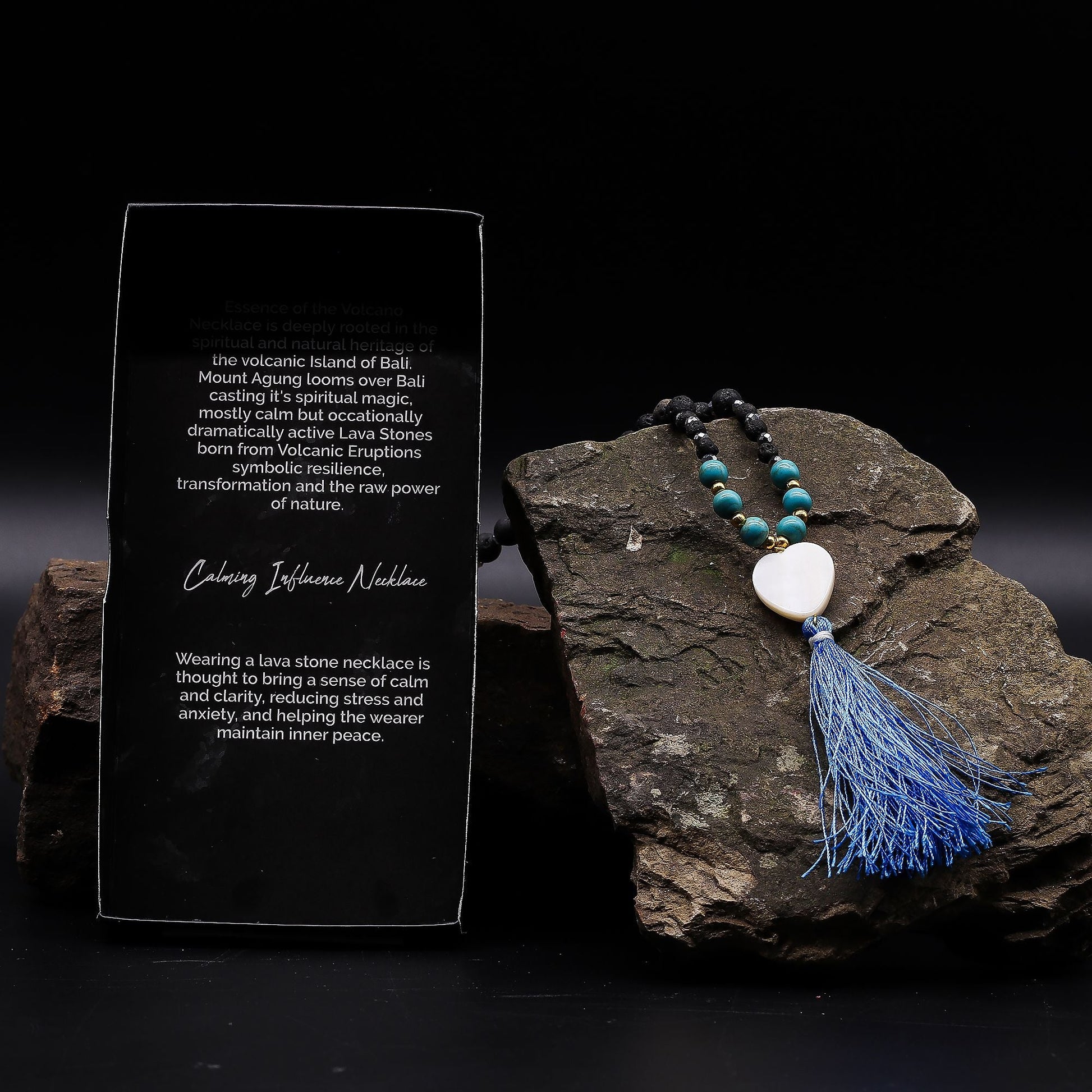 Essence of the Volcano Necklace – Calming Influence lava stone bead necklace with turquoise beads, white pendant and blue tassel on rock