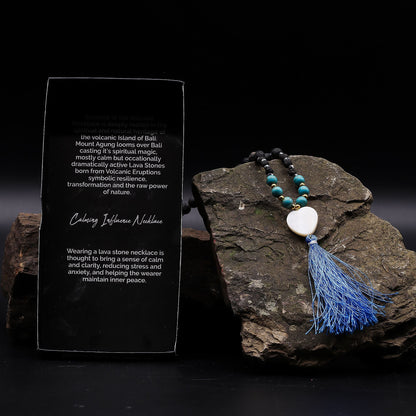 Essence of the Volcano Necklace – Calming Influence lava stone bead necklace with turquoise beads, white pendant and blue tassel on rock