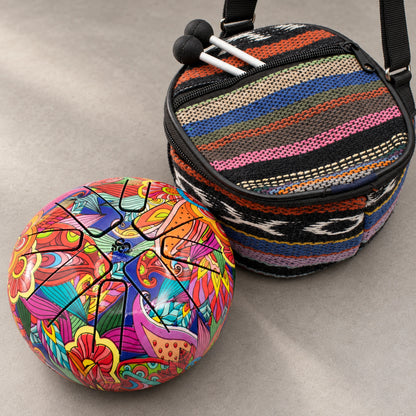 Colourfully patterned Happy Drum beside its woven carry bag, with two mallets at the open front pocket.