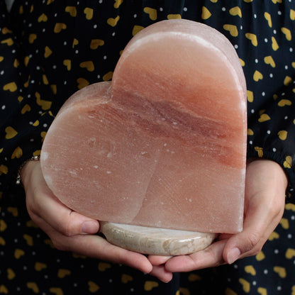 Hjärtformad Himalayasaltlampa handskuret rosa saltkristallhjärta på rund bas, hålls i händer, varm dekorativ accentbelysning