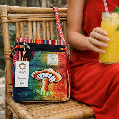 Hampa & Bomull Broderad Festväska – Orange Mushrooms crossbody-väska med orange svampbroderi på en stol bredvid en person som håller en drink