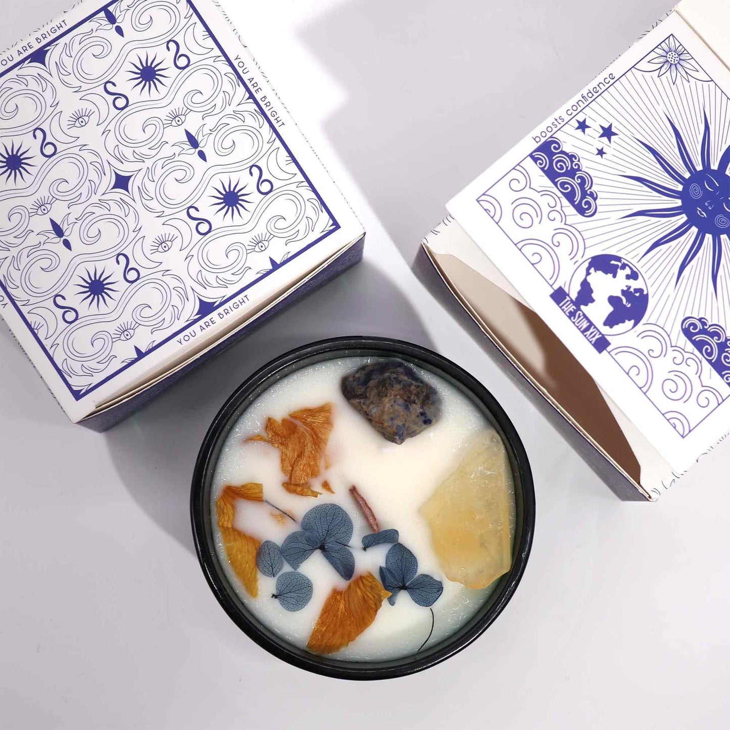 Alt Tag: Hope Hare Crystal Magic Flower Candle – The Sun med blomformad vax, kristaller och botaniska kronblad i svart plåtburk bredvid illustrerad ask