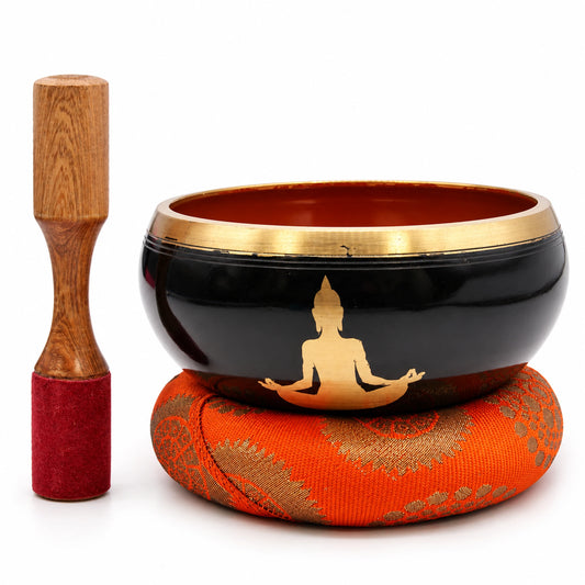 Black and orange Buddha singing bowl on an orange cushion, with its wooden striker standing beside it on white.