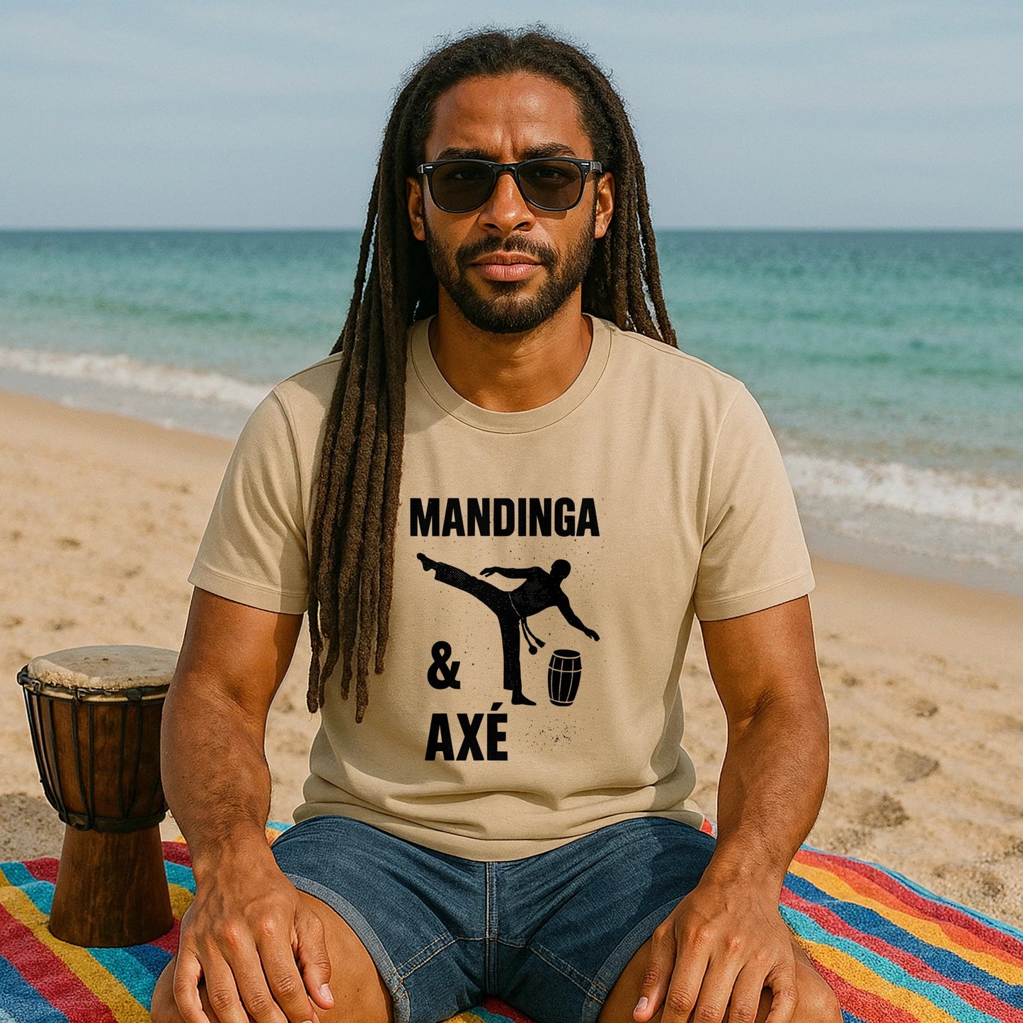 Alt Tag: Mandinga Axe Capoeira Premium Unisex T-Shirt beige, modelo sentado en la playa con gafas, estampado “Mandinga & Axé” y silueta capoeira