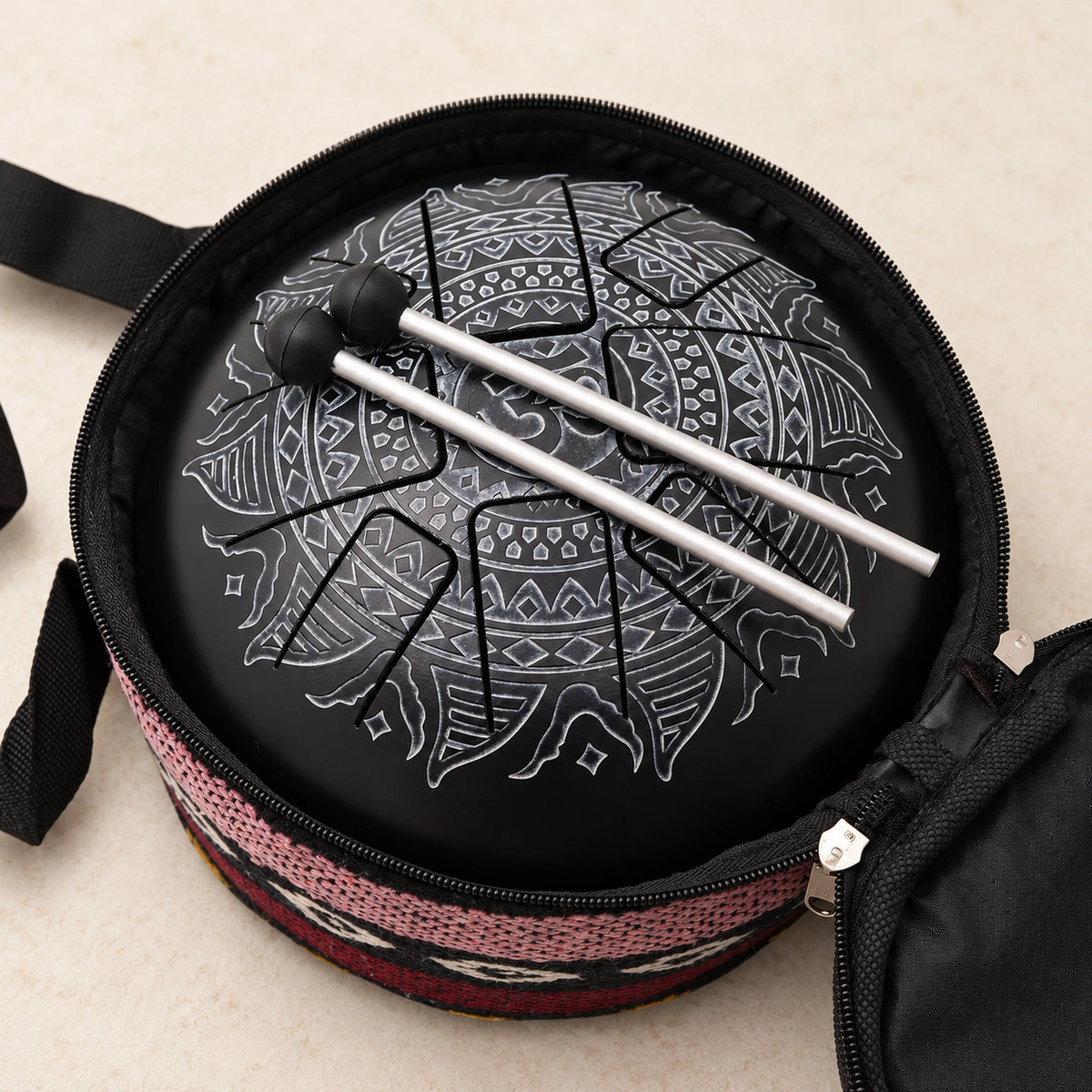 Black Om tongue drum with two mallets resting diagonally across it inside the open carrying bag with a patterned textile edge.