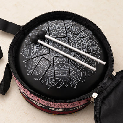 Black Om tongue drum with two mallets resting diagonally across it inside the open carrying bag with a patterned textile edge.