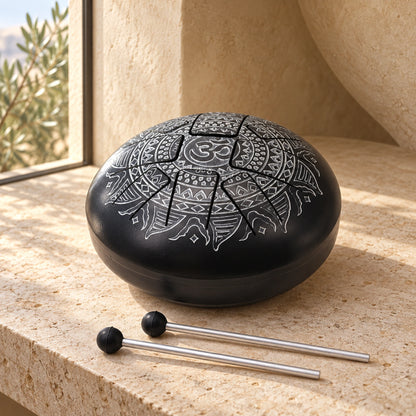 Black Om mandala tongue drum and two silver-coloured mallets on a pale stone console, beside a narrow window and curved sand-coloured plaster wall.
