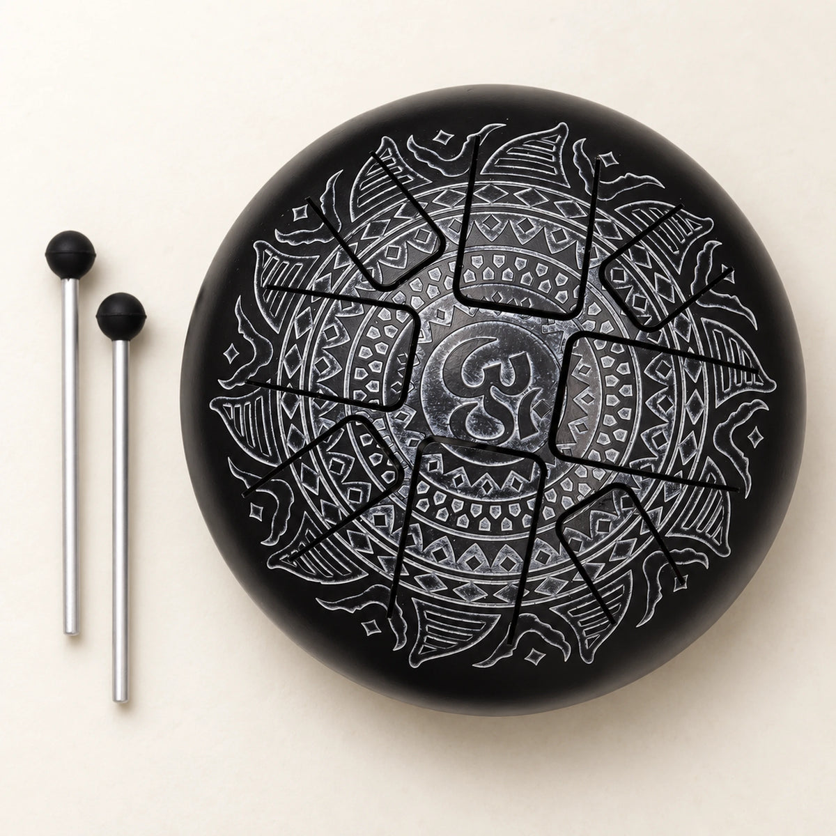 Black steel tongue drum with a silver-grey Om mandala and two mallets arranged to its left on an ivory background.
