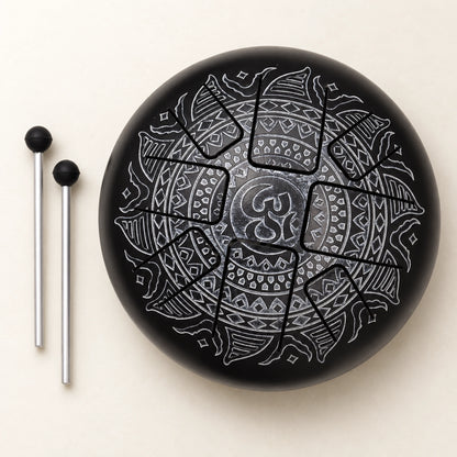 Black steel tongue drum with a silver-grey Om mandala and two mallets arranged to its left on an ivory background.