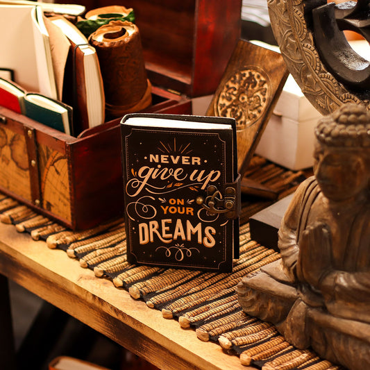 Alt Tag: Tryckt läderanteckningsbok – 17.8 × 12.7 CM Never Give Up On Your Dreams på ett skrivbord i trä med böcker och dekorationer
