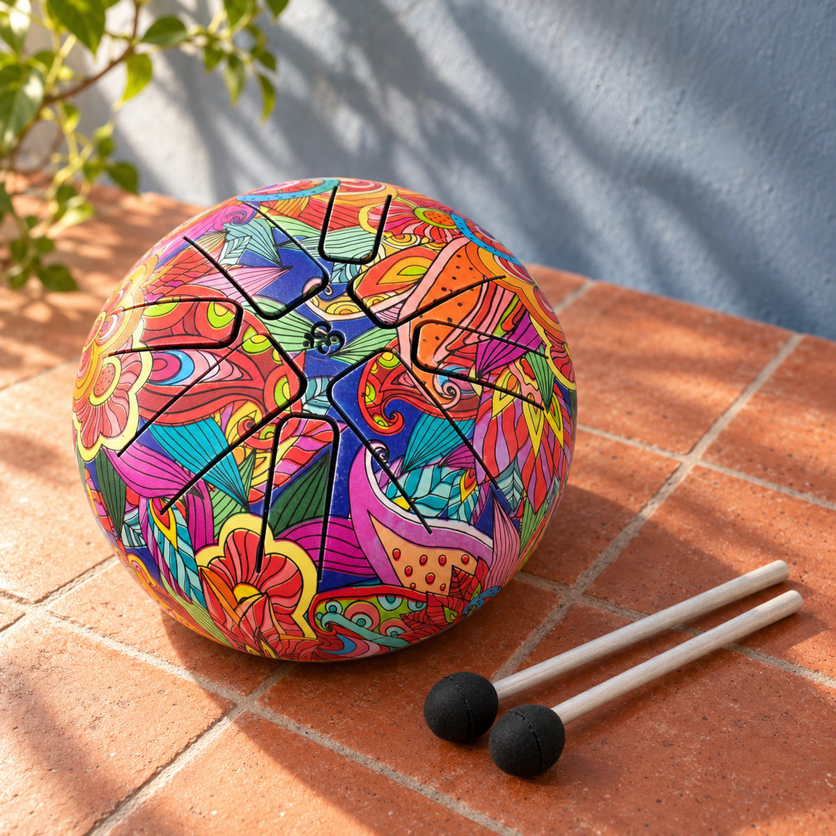 Colourfully patterned Happy Drum and two black-headed mallets on terracotta tiles, against a blue-grey wall with soft leaf shadows.