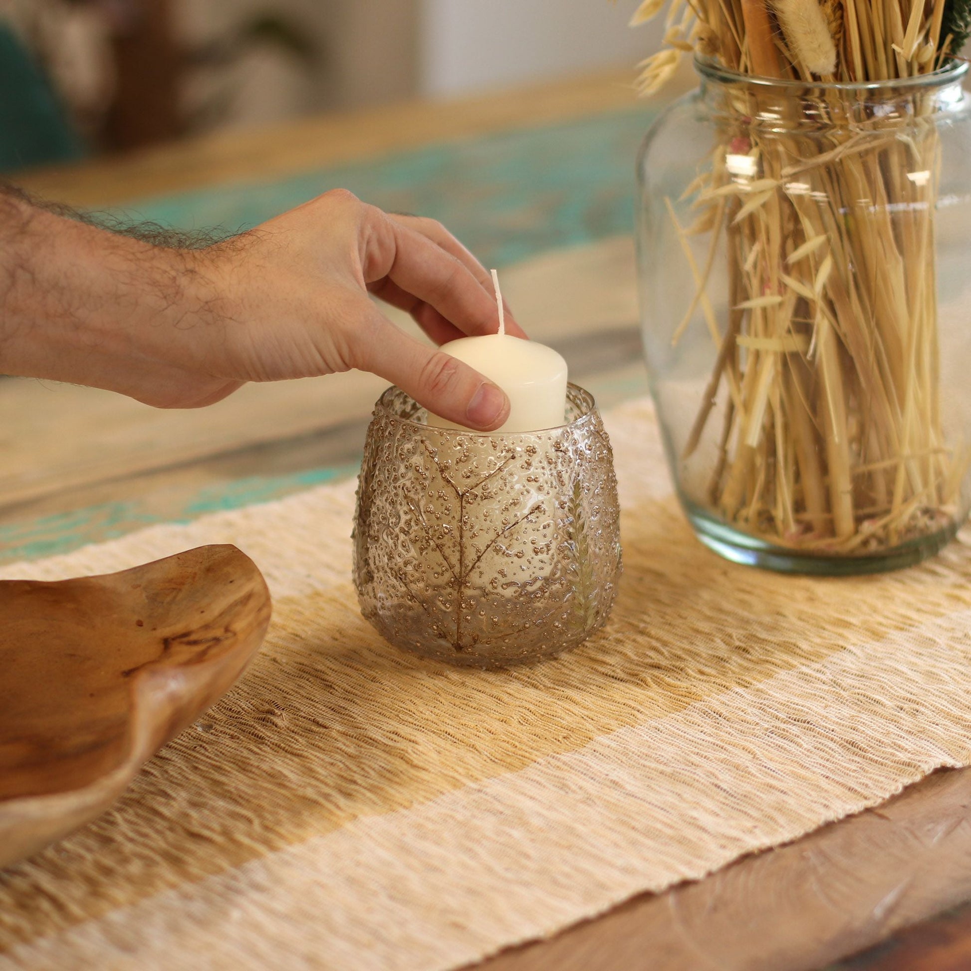 Alt Tag: Portavelas de vidrio reciclado – 10x9 CM con diseño de hoja de helecho, mano colocando una vela tipo tealight sobre decoración de mesa rústica