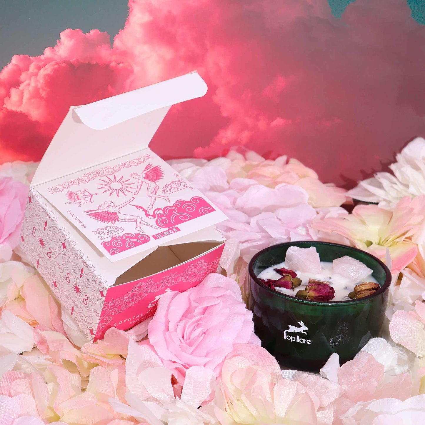 Alt Tag: Soy Wax Crystal Candle – The Lovers en vaso verde con cristales y pétalos de rosa, junto a caja rosa sobre flores