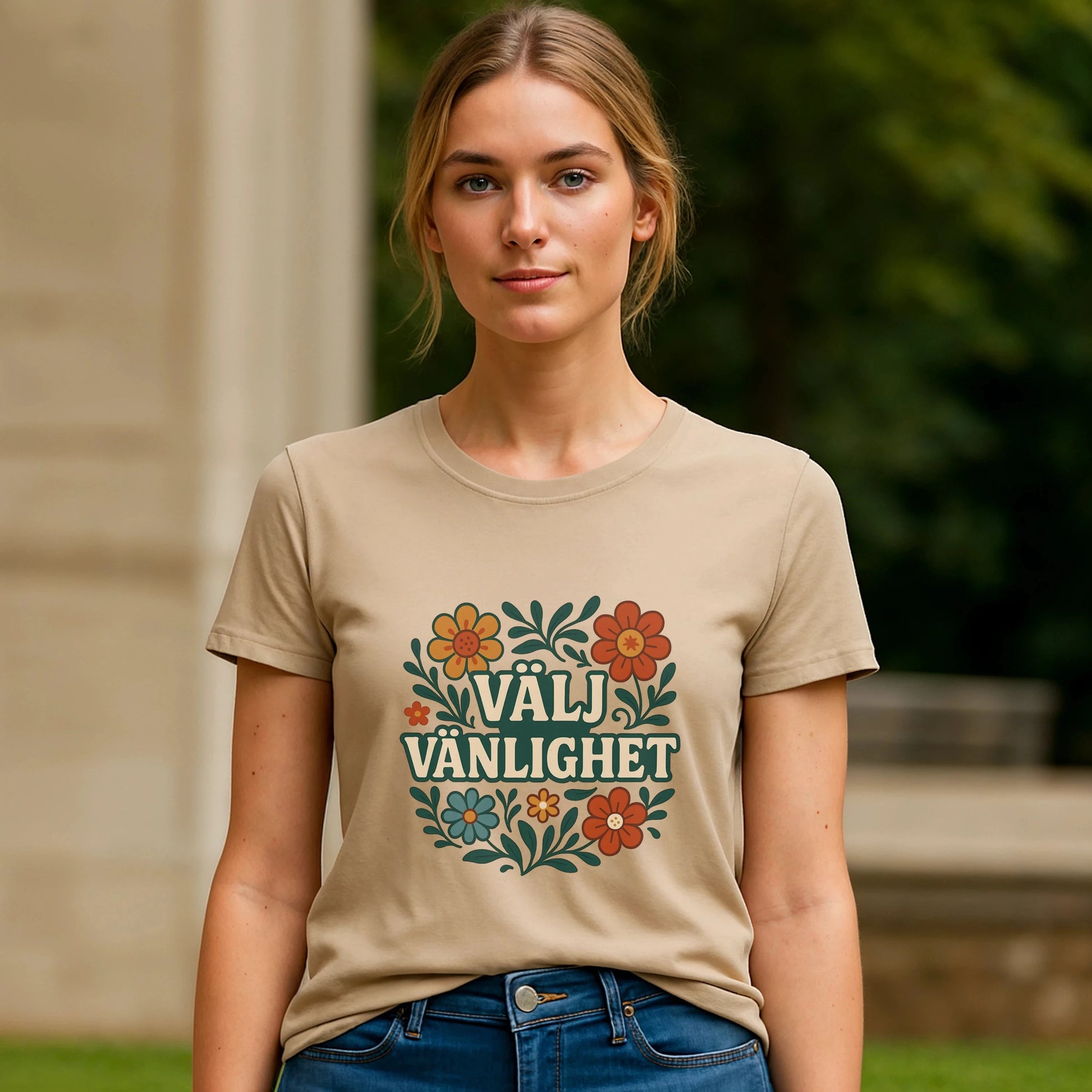 Alt Tagg: Modell bär Välj Vänlighet T-shirt i 100% ekologisk bomull i beige med blommigt "Välj Vänlighet"-tryck