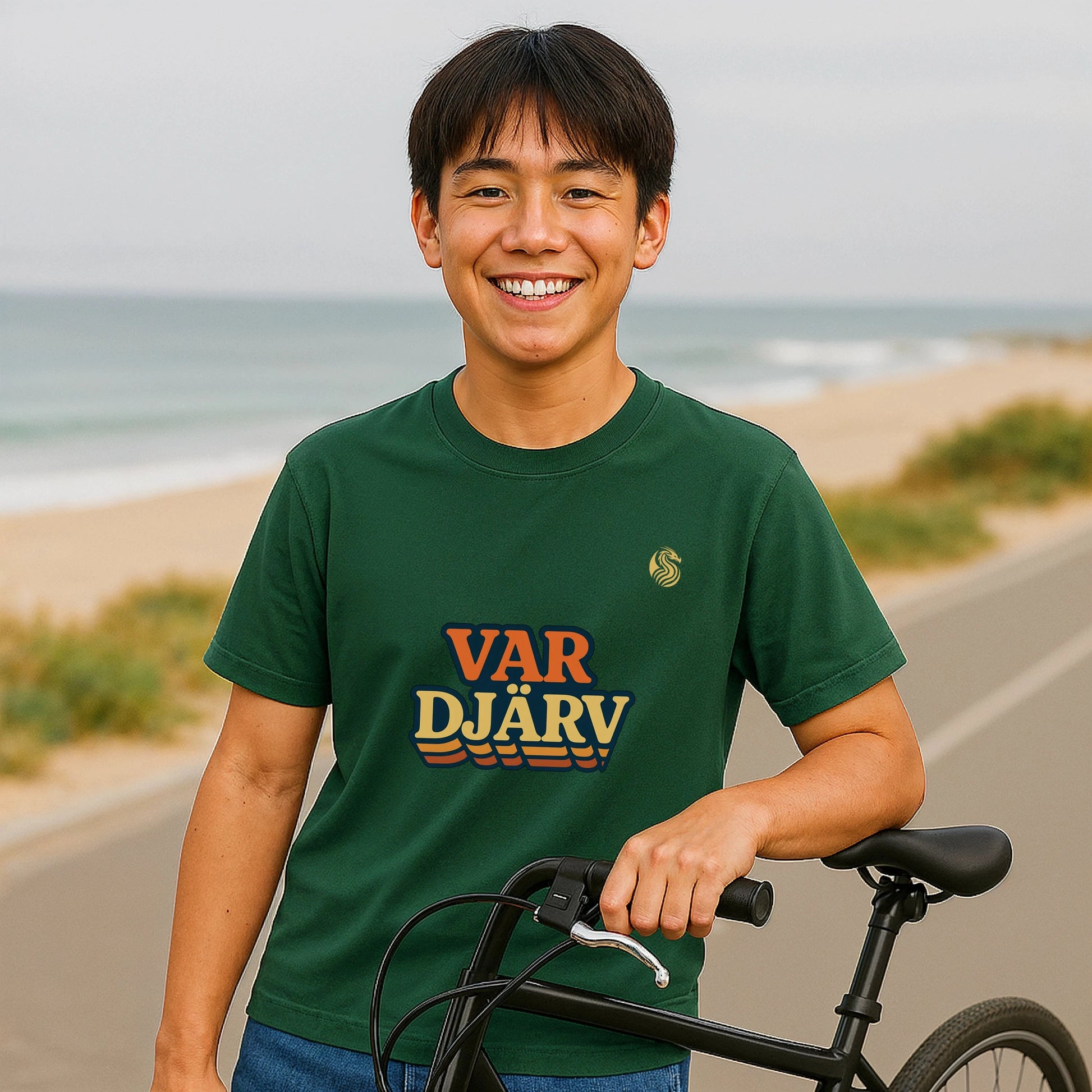 Var Djärv Ungdom 100% Organic Cotton T-Shirt on smiling person wearing green graphic tee, holding bicycle by a beach road