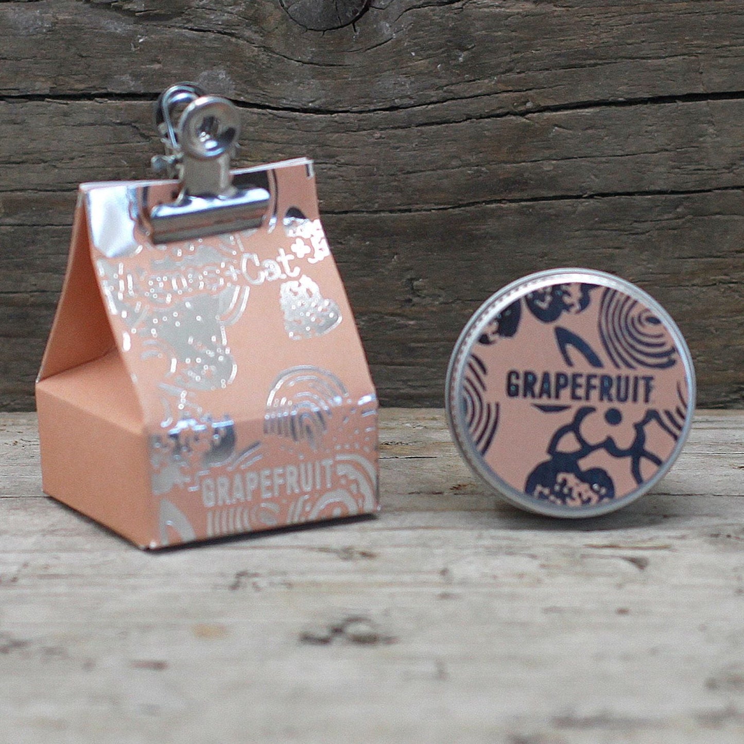 Alt Tag: Vegan Grapefruit Lip Balm – 12G in tin container with grapefruit label beside peach gift box on rustic wooden background